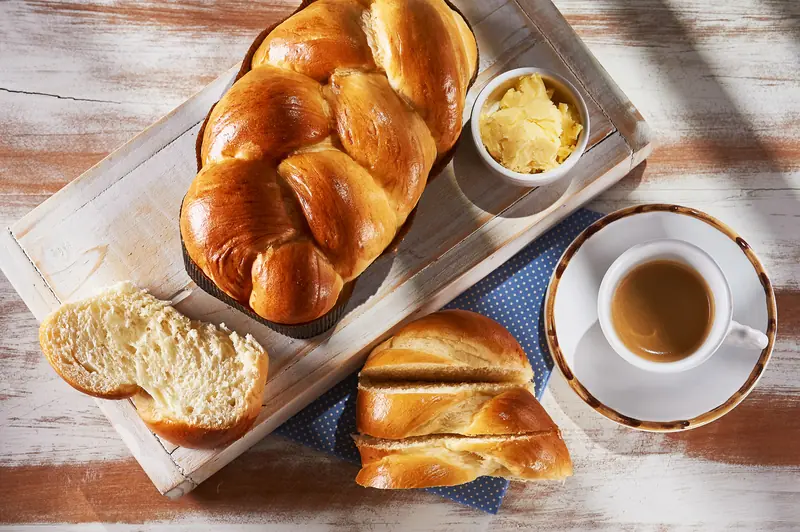 Mesa de café da manhã vista de cima: trança de brioche dourada, fatias macias, manteiga e uma xícara de café sobre madeira clara