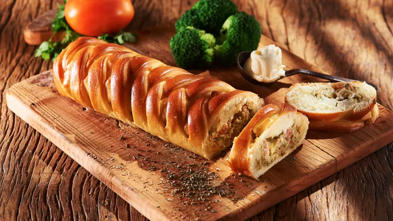 Pão trançado recheado de frango e legumes, fatiado sobre tábua rústica com brócolis fresco, tomate e uma colherada de requeijão