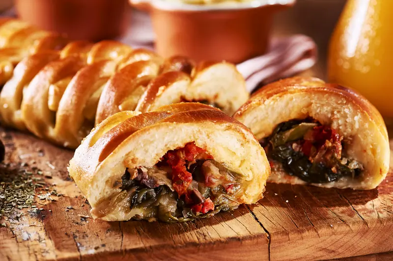 Fatias de pão trançado com recheio generoso de escarola refogada e tomate seco, sobre tábua rústica