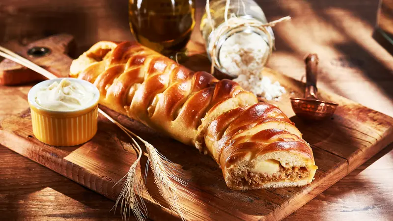 Pão trançado dourado recheado de frango desfiado com requeijão cremoso, com uma fatia aberta sobre tábua rústica, ao lado de azeite, farinha e espigas de trigo
