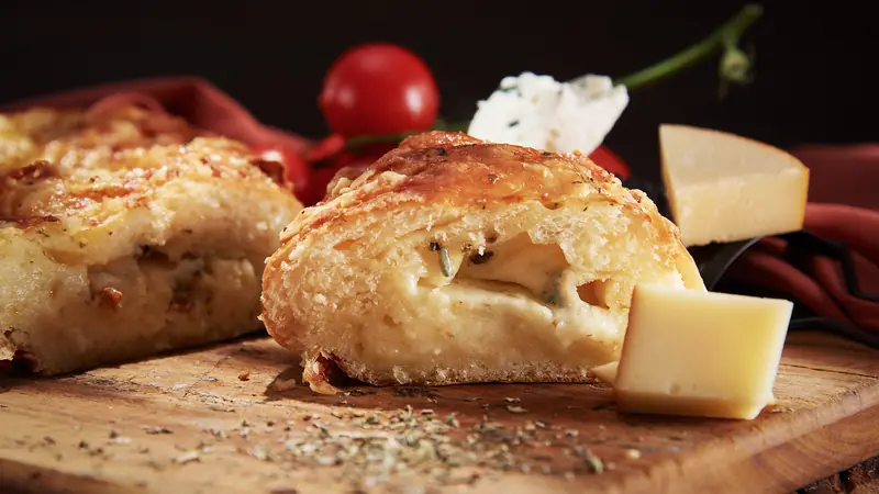Pão recheado de queijos com casca gratinada e interior cremoso, ao lado de lascas de queijo e tomates-cereja sobre fundo escuro